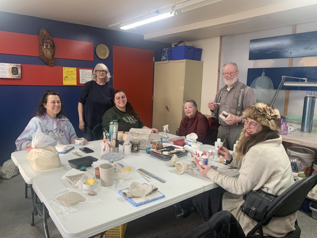 Six people around a work table in Kathleen's studio, hand-building with clay — rolled slabs, tools and pots in progress in front of them