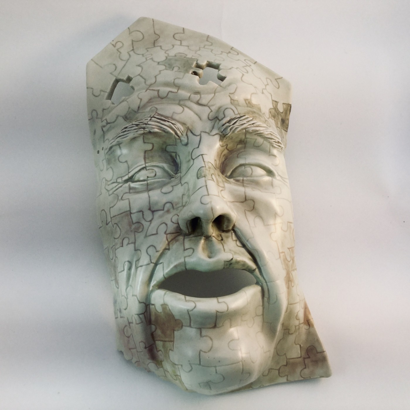 Life-size ceramic face mask with an open mouth, its entire surface incised with interlocking jigsaw-piece lines and a few pieces missing from the forehead