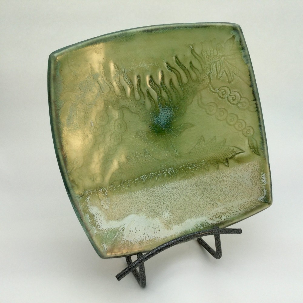 Square plate on a wire stand, glazed in mottled green and gold, with a fern frond and leaves impressed into the clay