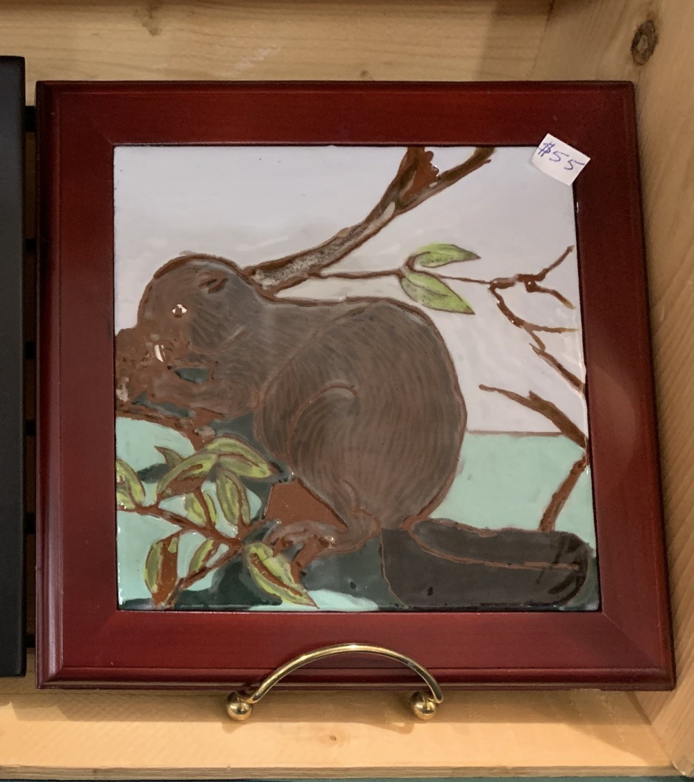 Glazed tile of a dark woodrat on a branch among green leaves, in a red wooden frame