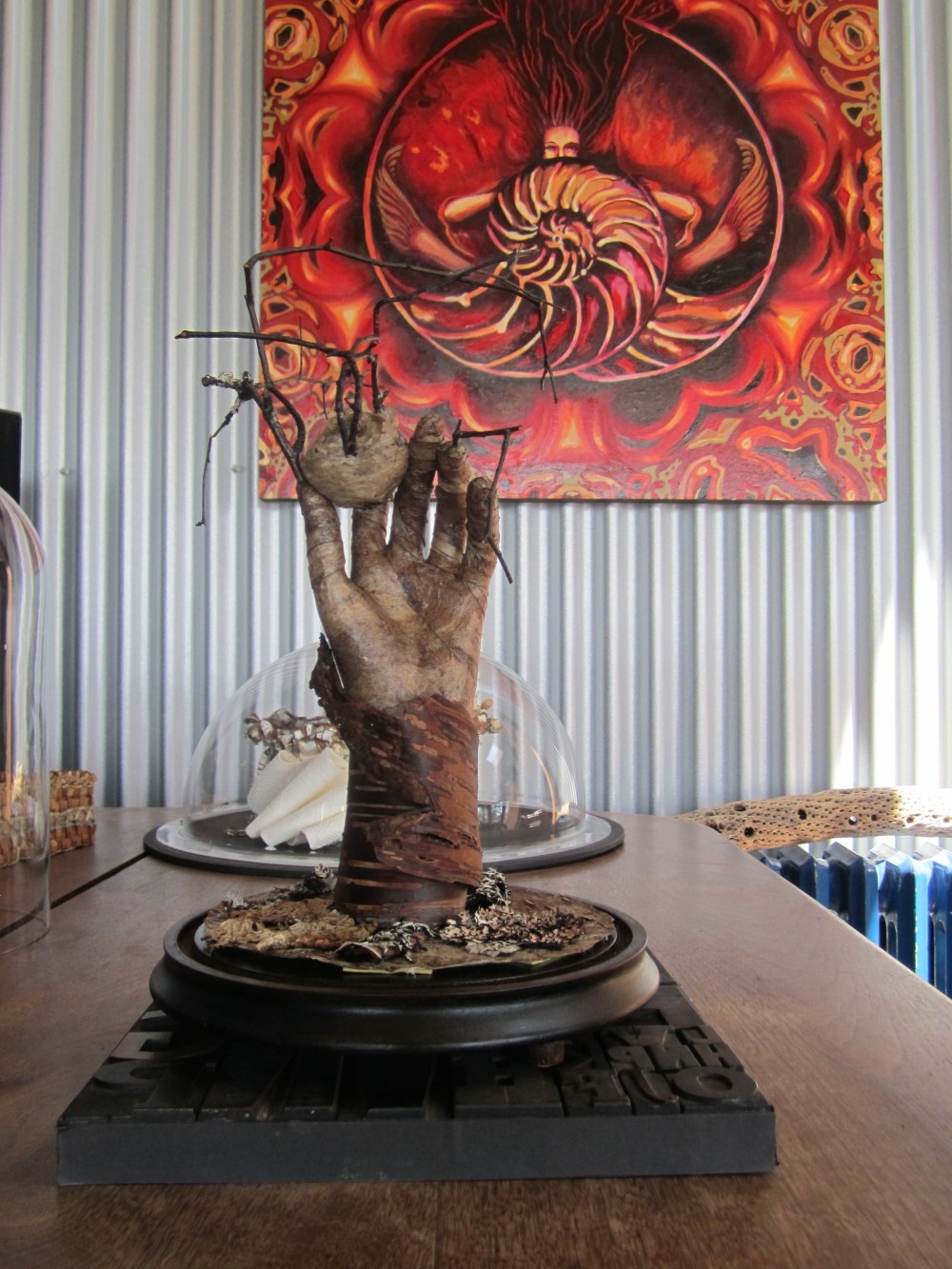 Sculpture of a weathered hand reaching upward from a bark base, holding a wasp nest and bare twigs, mounted on a dark bell jar base
