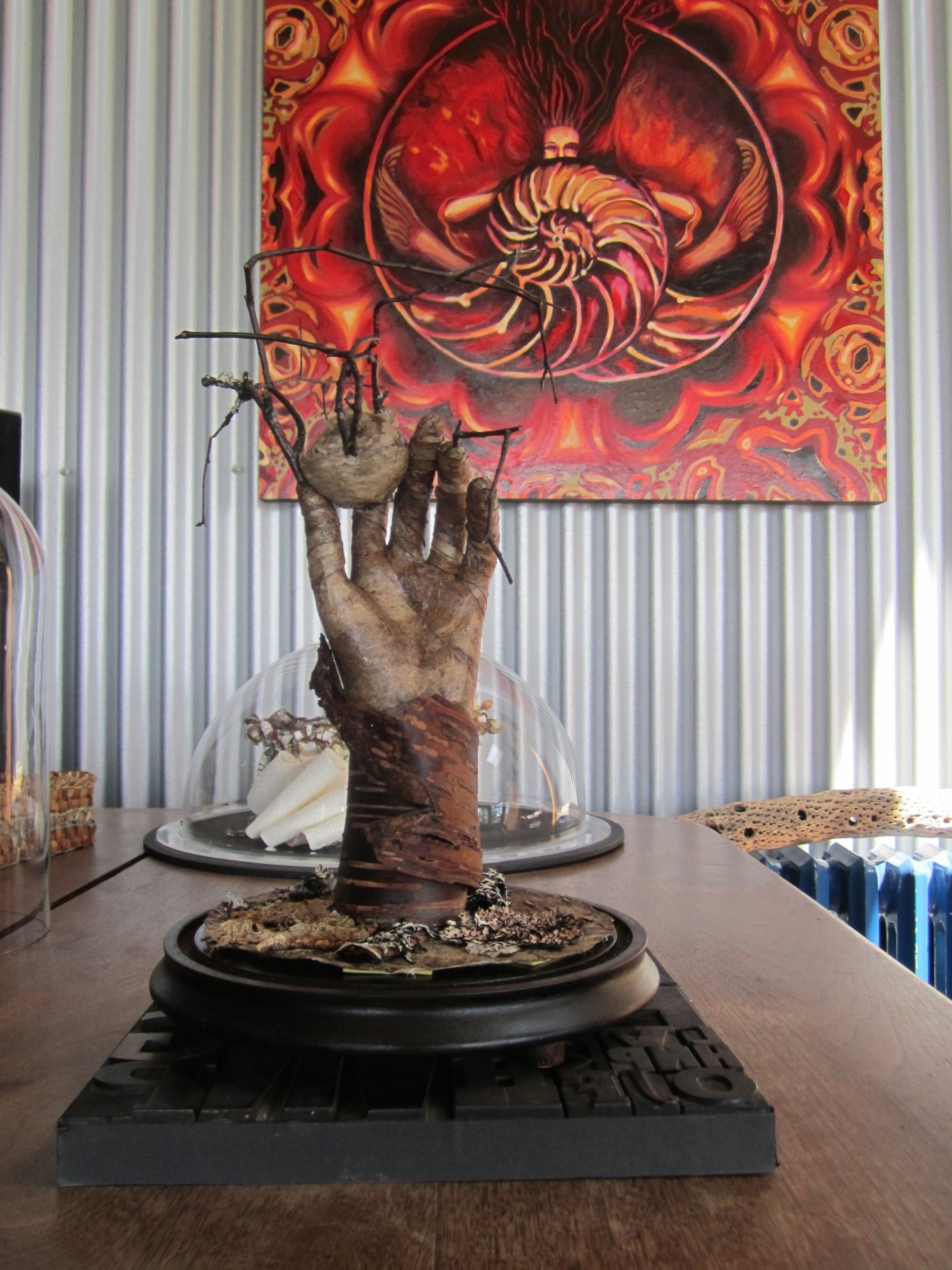 Sculpture of a weathered bark covered hand reaching upward from a bark base, holding a wasp nest and bare twigs, mounted on a dark bell jar base