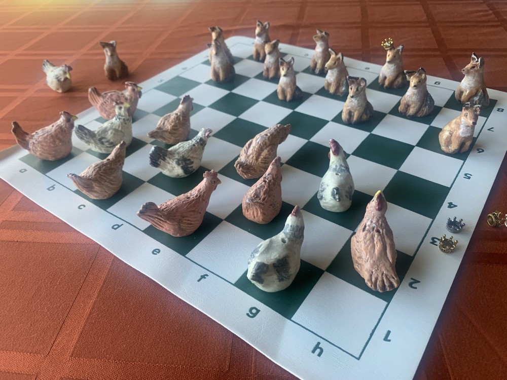 A chess set of small clay figures laid out on a board — hens on one side, foxes on the other, the kings and queens wearing tiny gold crowns