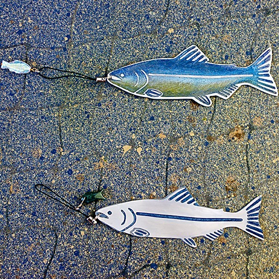 Two flat etched metal salmon ornaments on paving stone, one glazed blue and green, one left white with blue linework