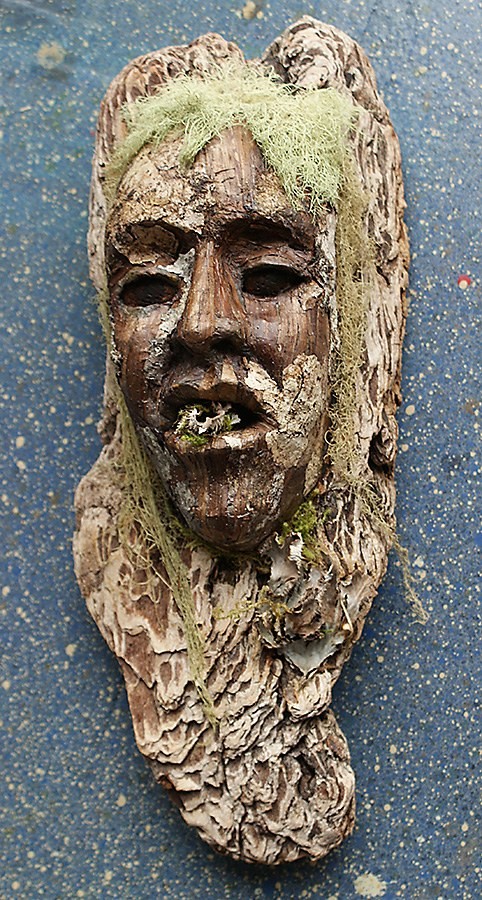 Narrow bark mask with a carved face, pale green lichen falling either side like long hair, and moss in the open mouth