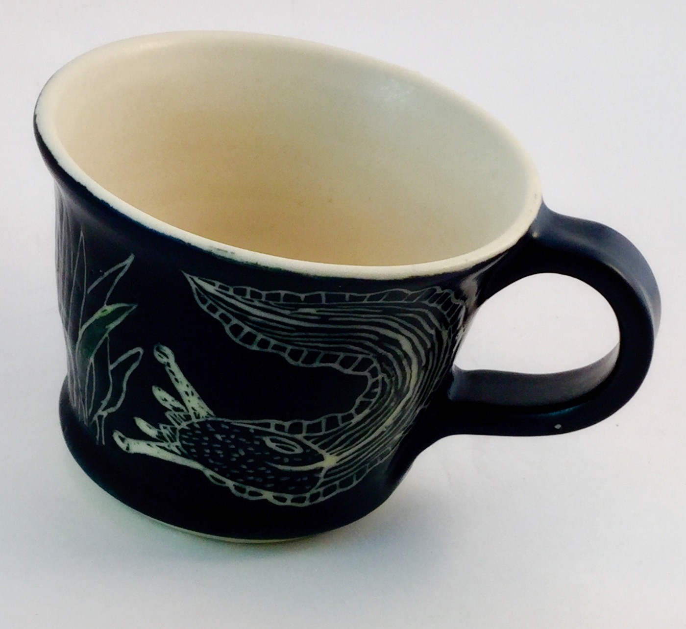 Black stoneware mug with a banana slug and leaves drawn through the glaze in white sgraffito, cream glazed interior
