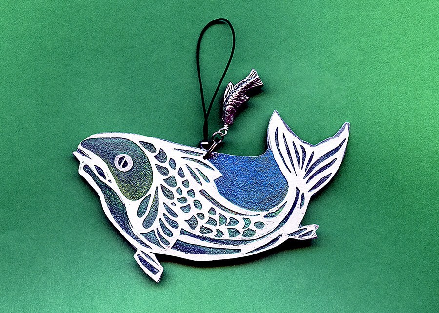 A single etched metal fish ornament with scales and fins cut through the surface, glazed blue and green, hanging from a cord with a small pewter fish charm