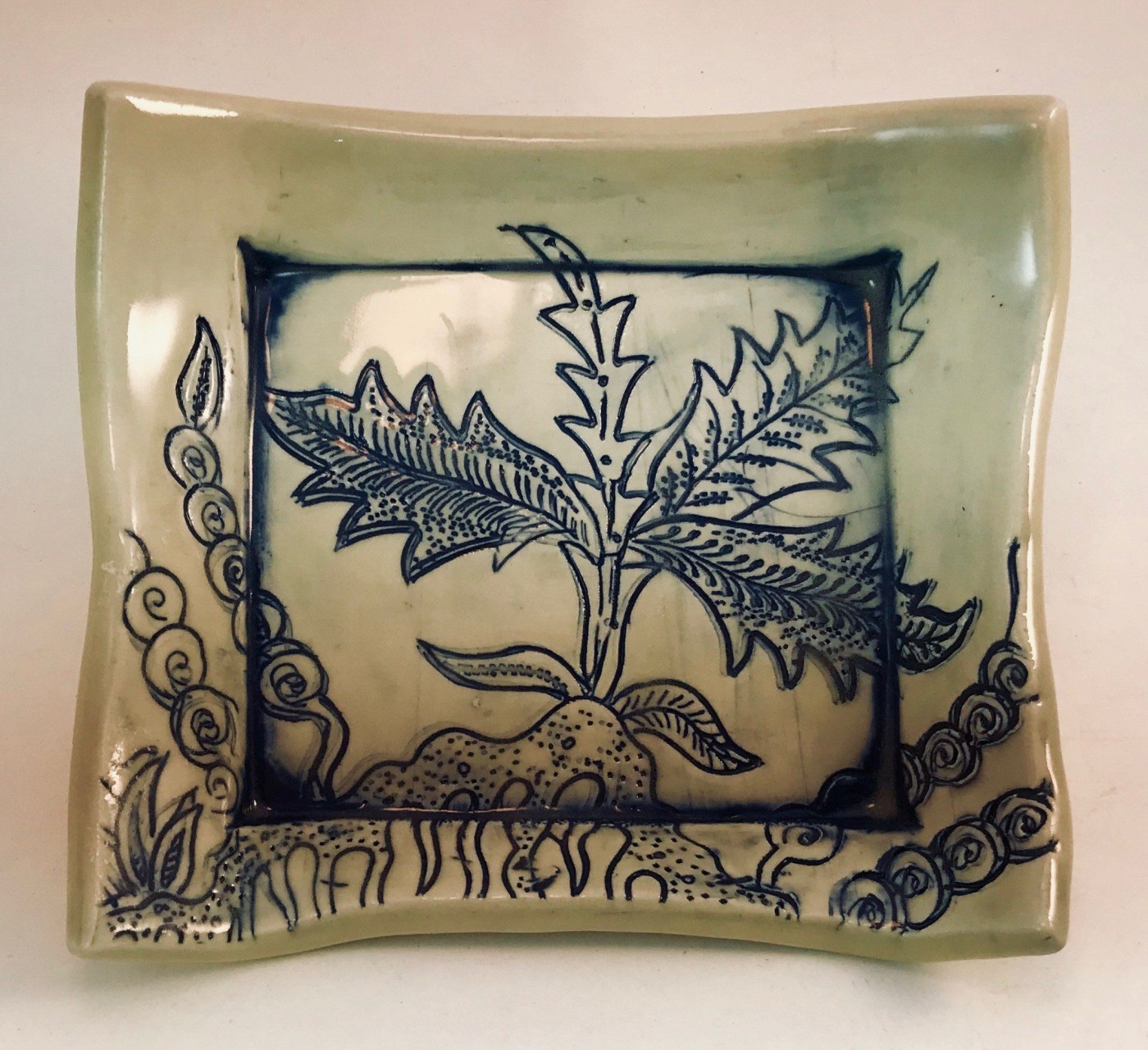 Shallow square celadon dish with a dandelion-like plant drawn in cobalt line, framed by a border of spirals and grasses