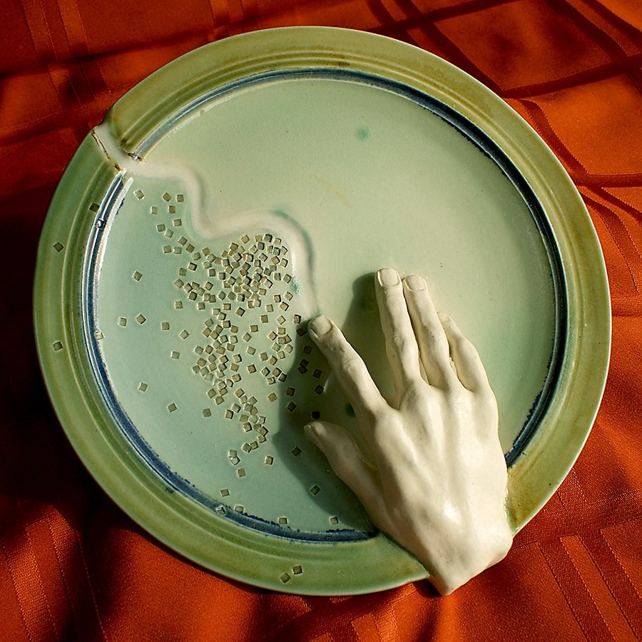 Celadon plate with a life-cast porcelain hand resting on the rim and a scatter of small clay squares curving across the well