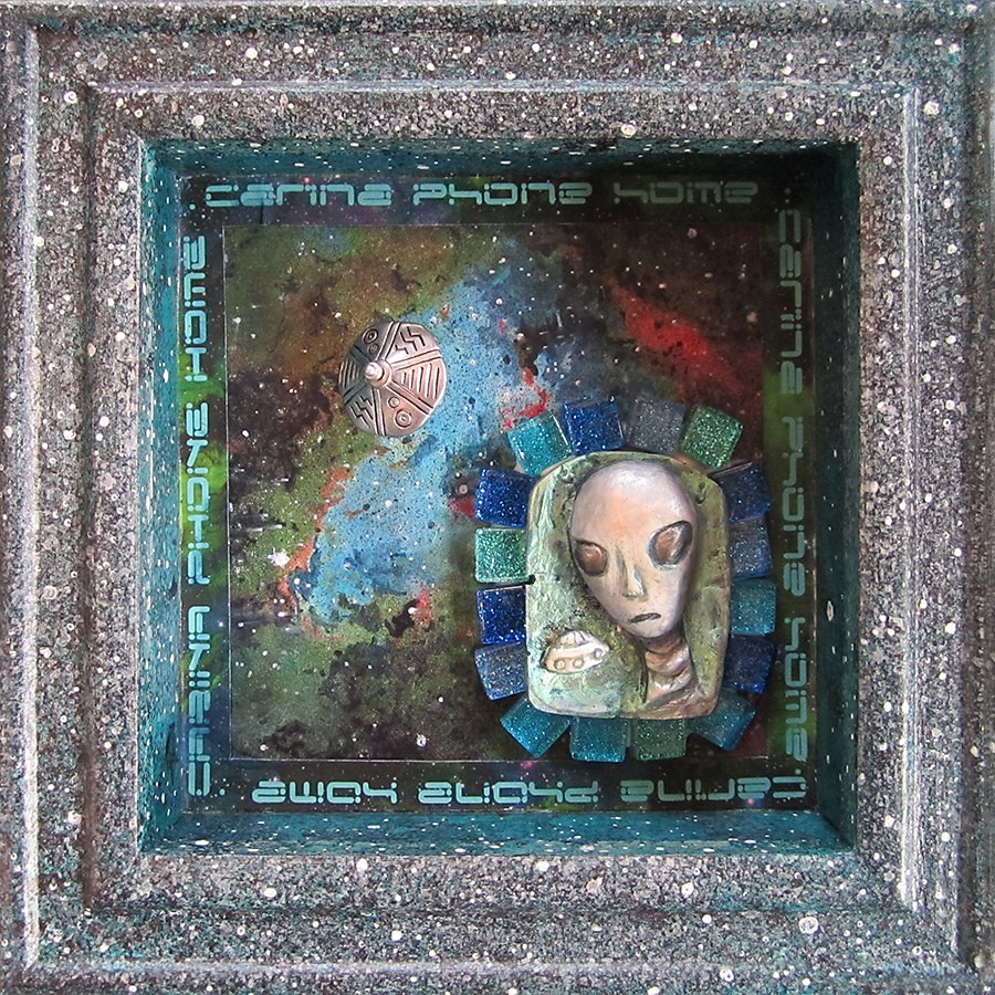 Shadowbox assemblage with a pale clay alien face on blue and green glass tiles against a painted nebula, framed in speckled grey with lettering around the inner edge