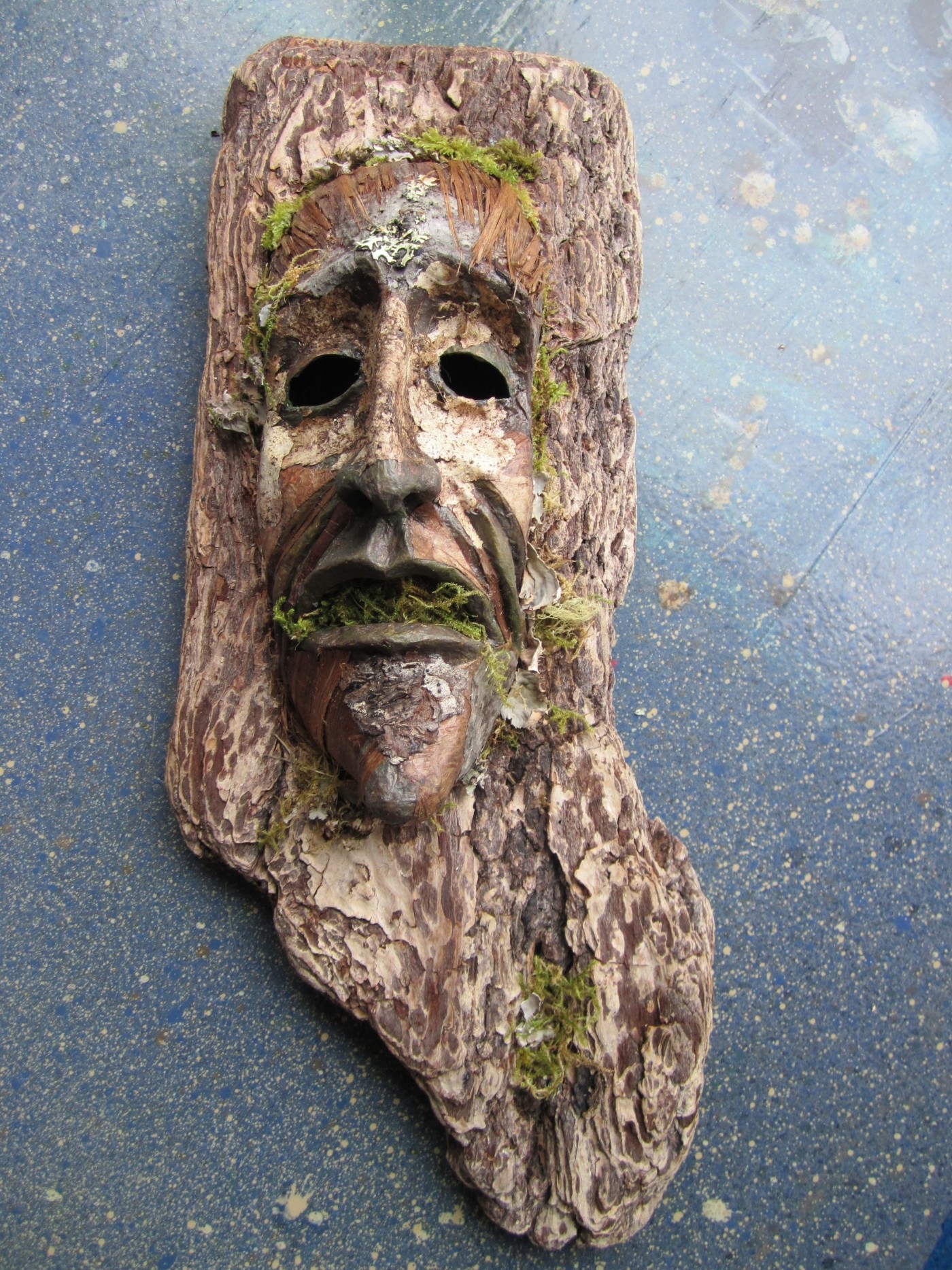 Mask carved from a slab of cedar bark, with a long face, moss  and lichen for eyebrows and beard, and eyes made from almonds.