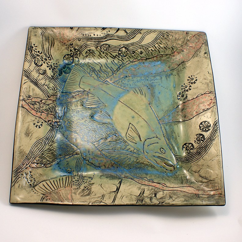 Square platter glazed in sea green and blue, a large salmon carved in relief across the surface among incised waves and barnacles