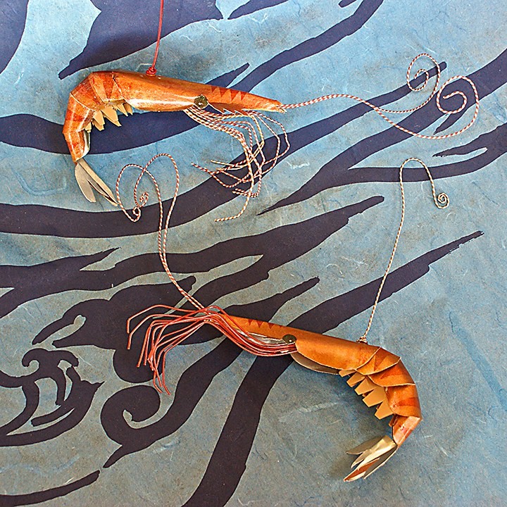 Two prawns made from painted aluminum sheet and wire, their long antennae curling, laid on indigo paper printed with a wave pattern